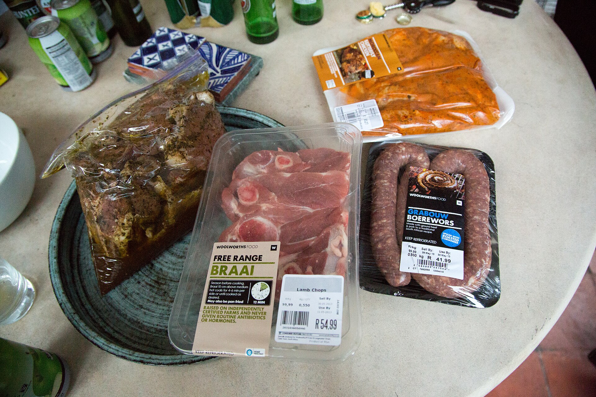 Packages of raw meat on a table: a marinated beef roast, lamb chops, boerewors and chicken wings.