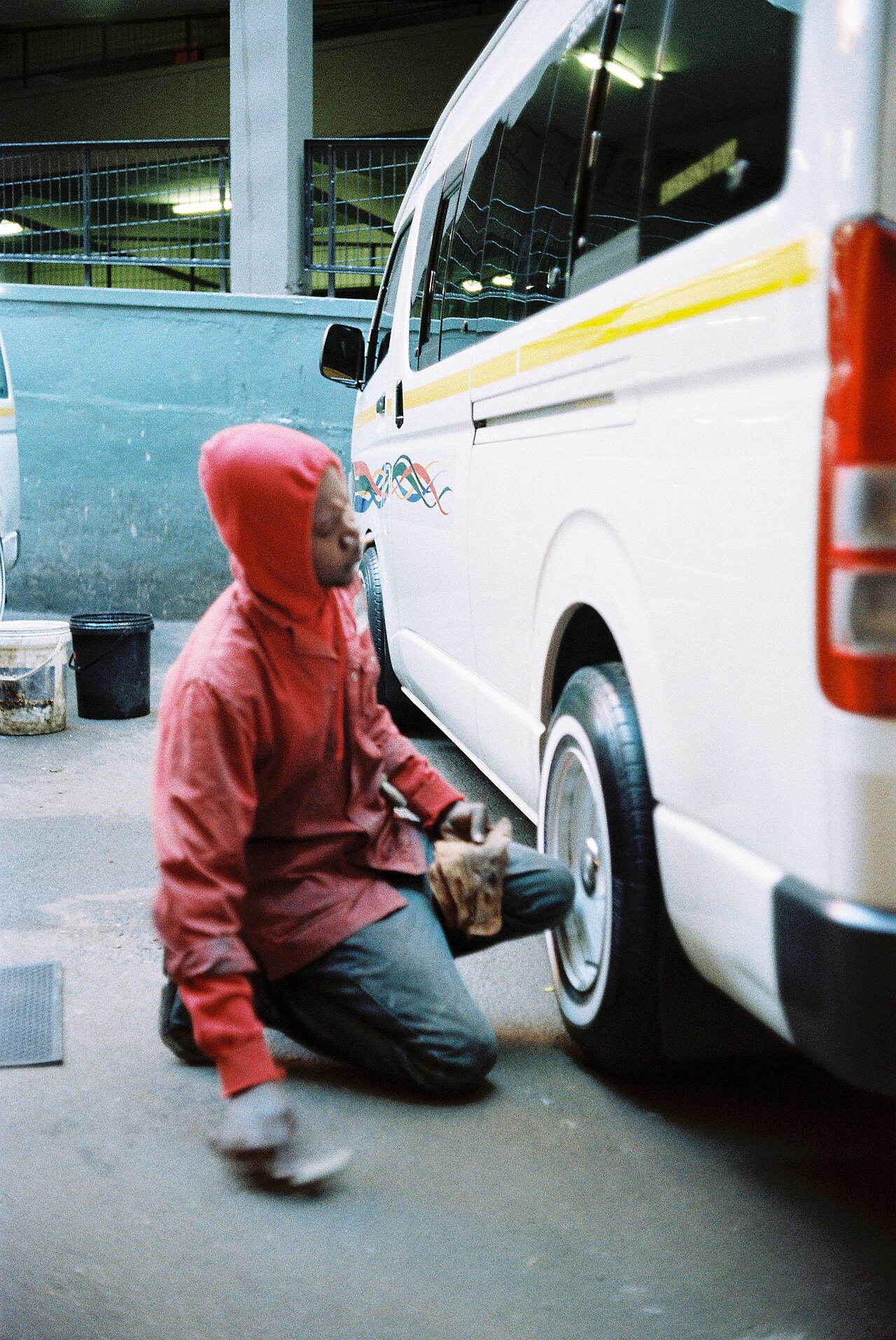 A young person in a red hoodie kneeling beside a white van, checking a tyre with a small device.
