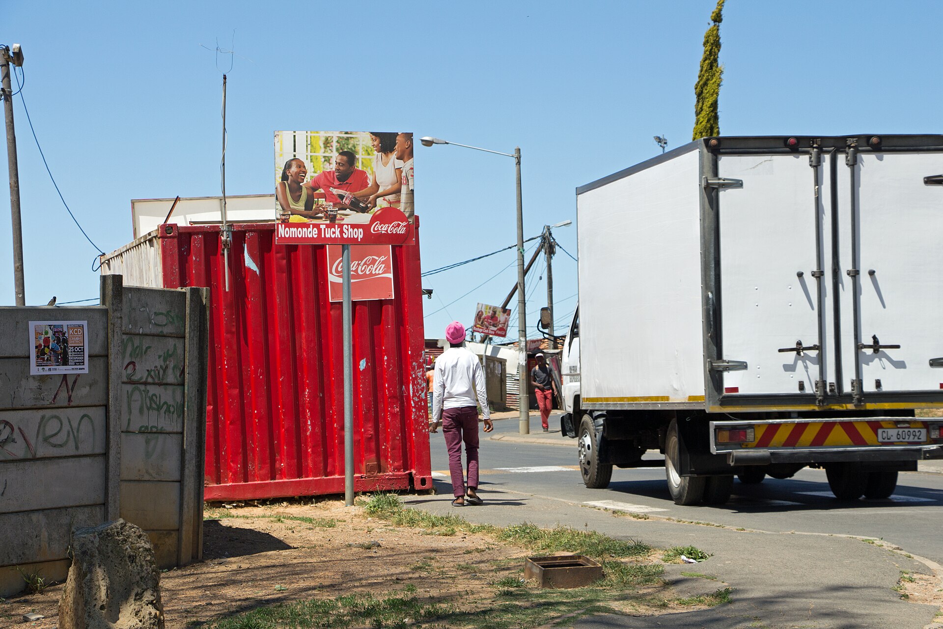 A red shipping container used as a tuck shop, with a large Coca-Cola advertisement on its side and a delivery truck nearby.